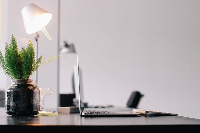 mała biała lampka na ciemnym biurku z notebookiem i roślina w słoiku