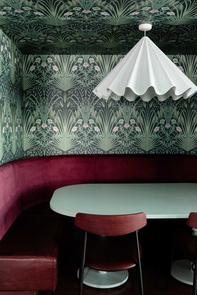wnętrze lokalu usługowego projektu Kingston Lafferty Design zielony stolik z czerwonym siedziskiem pod białą lampą na tle ściany z zieloną roślinną tapetą