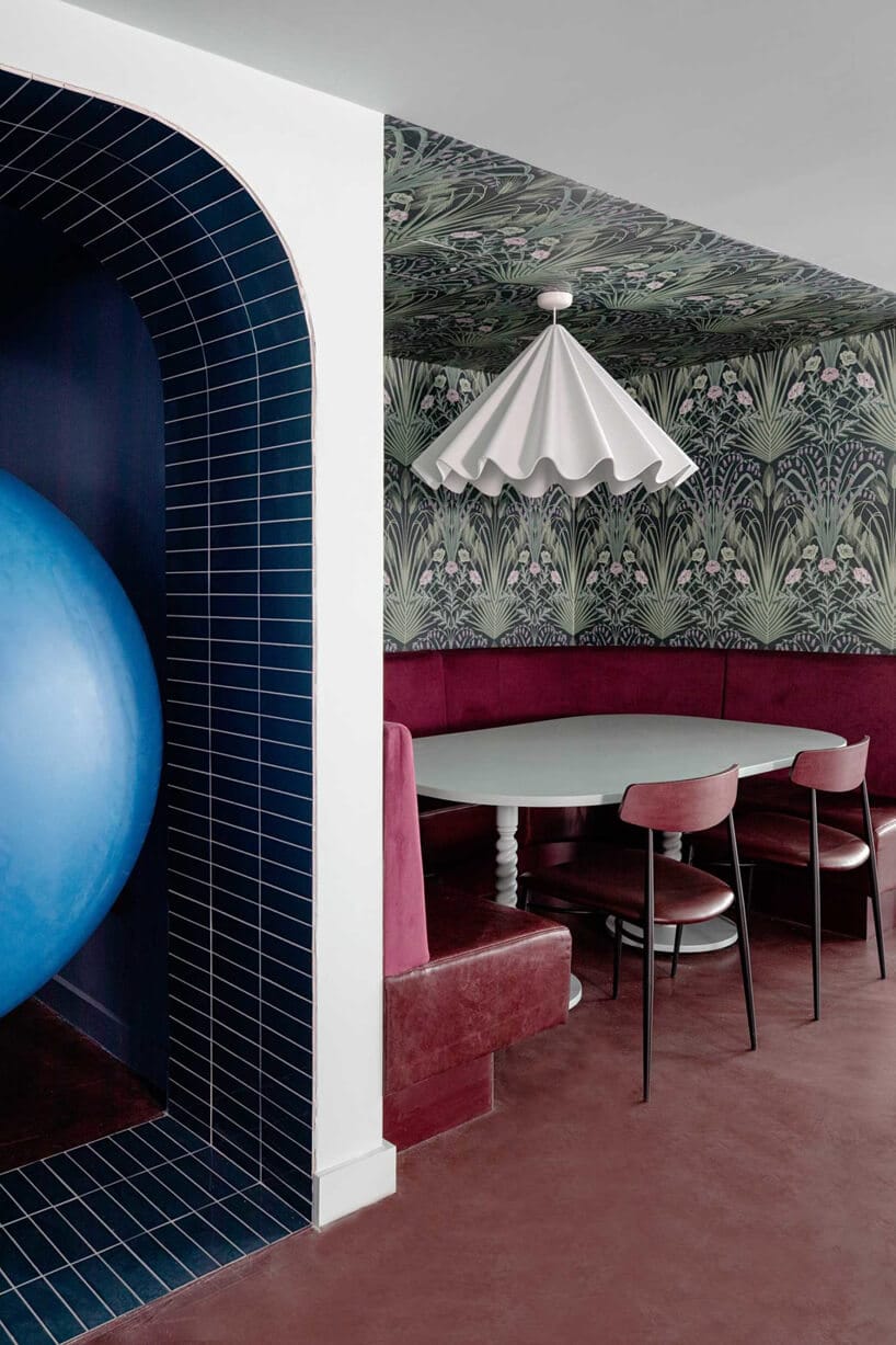 wnętrze lokalu usługowego projektu Kingston Lafferty Design zielony stolik z czerwonym siedziskiem pod białą lampą na tle ściany z zieloną roślinną tapetą obok niebieskiej wnętki
