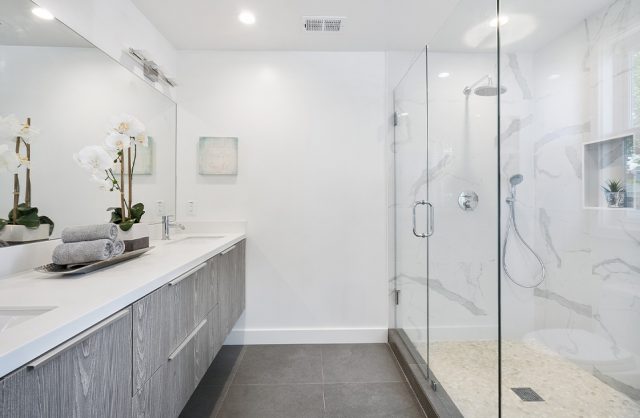 biała łazienka z długim natryskiem za szklana ścianą na przeciwko podwieszonych szarych szafek z białym blatem