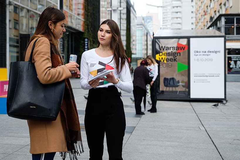 hostessa w białej bluzce rozmawiająca z kobieta w płaszczu obok reklamy Warsaw Design Week 2019