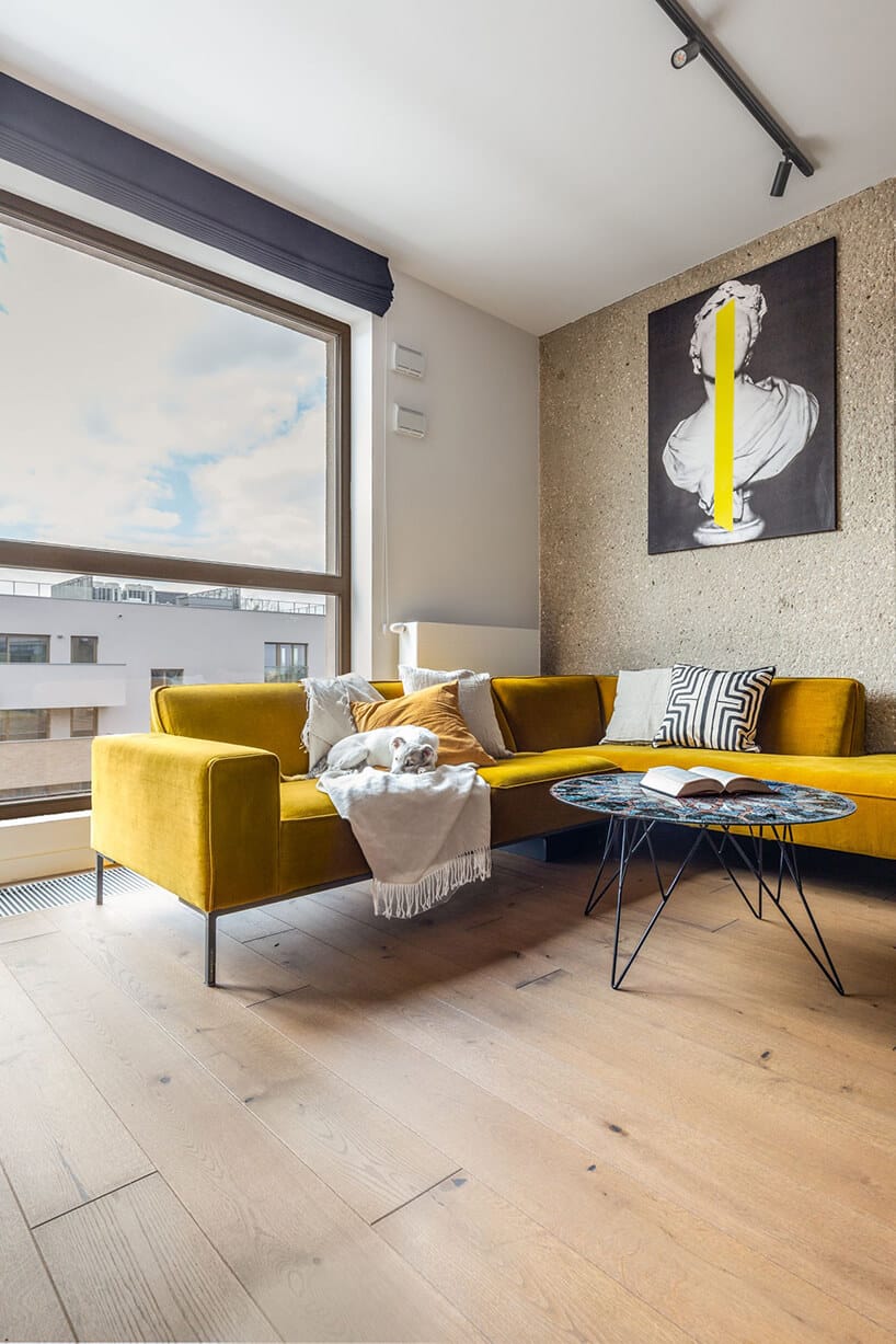 wnętrze w stylu hygge narożna żółta sofa przy okrągłym stoliku na tle dużego panoramicznego okna