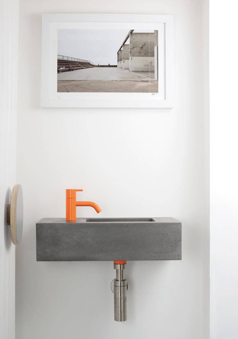 szara betonowa umywalka z pomarańczową baterią w białej łazience