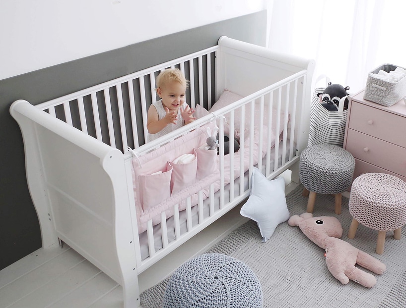 białe ładne drewniane wysokie łóżeczko od Woodies Safe Dreams dla dziecka z różową pościelą i małą dziewczynką w środku