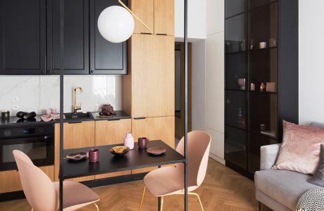 kawalerka od ZAZA Studio czarno brązowy aneksy kuchenny jako tło dla czarnego stolika z dwoma różowymi krzesłami ze złotymi nogami
