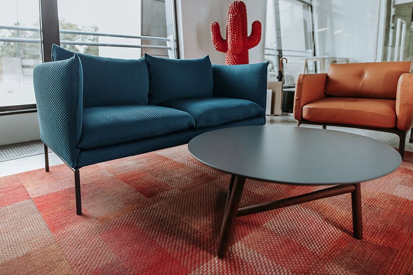 małą niebieska sofa na wysokich nogach obok pomarańczowego fotela przy okrągłym stoliku na czerwonym dywanie
