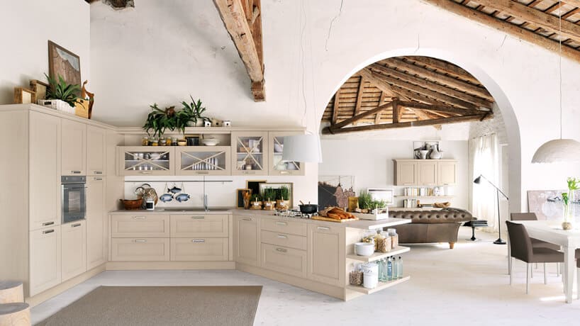 przestronna biała kuchnia w stylu prowansalskim z kremowymi szafkami z wyspą pod skosem pod drewnianymi starymi filarami na tle wejścia do salonu