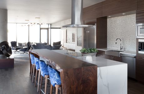 wnętrze penthouse'u od Benning Design Construction brązowa kuchnia z białą wyspą z kamienia i wysokim ciemnym drewnianym stołem z czterema krzesłami z niebieskim oparciem