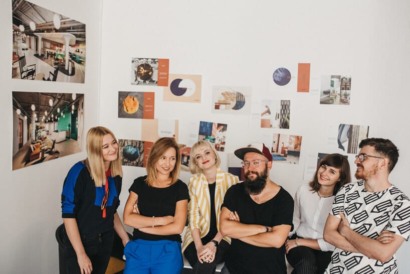 sześć osób opartych o biurko na tle ściany z projektami