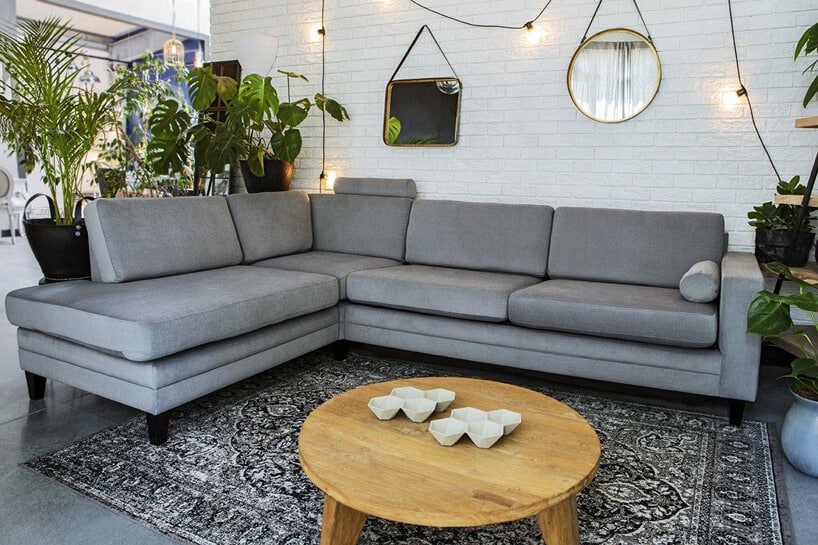 szara sofa narożna Focus od 9Design na krótkich czarnych nóżkach przy drewnianym okrągłym stoliku na szarej podłodze na tle białej ceglanej ściany