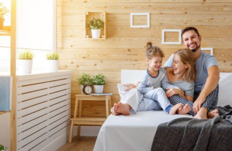 szczęśliwa rodzina siedząca na dużym łóżku na tle drewnianej ściany ozdobionej ramkami i kwiatami w białych doniczkach