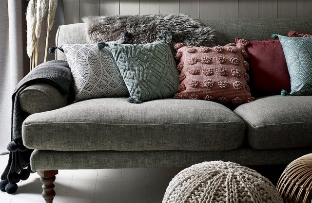 szara sofa na drewnianych krótkich nogach z pięcioma poduszkami o różnym kolorze i fakturze materiału