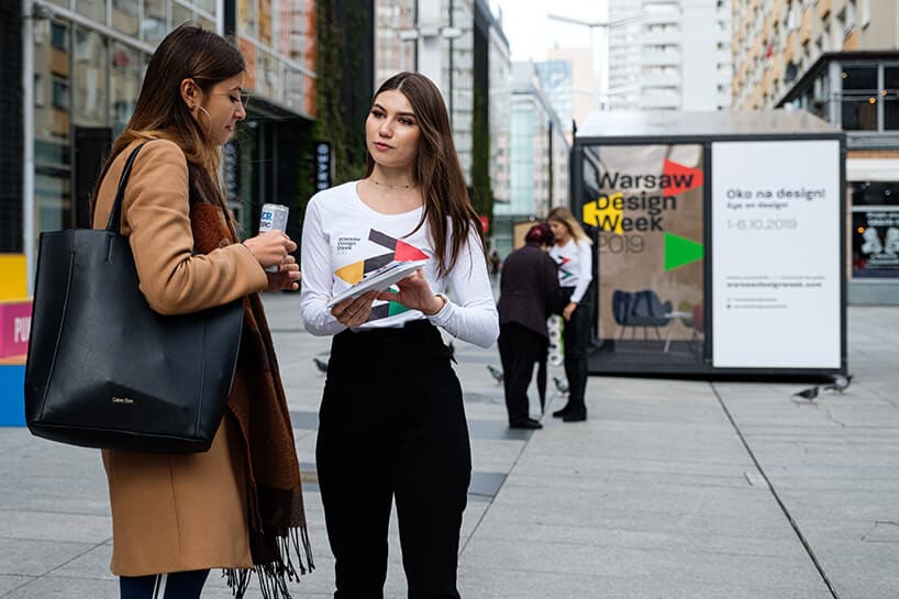 hostessa w białej bluzce rozmawiająca z kobieta w płaszczu obok reklamy Warsaw Design Week 2019
