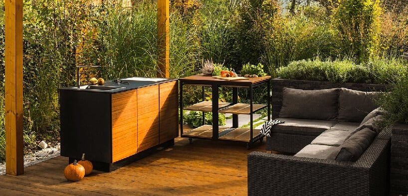 drewniane patio z ciemną szafką ze zlewem przy czarnej szafce na kółkach na tle wysokich roslin w ogrodzie