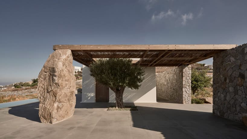 biały kamień podtrzymujący drewniany dach przy wejściu do willi z małym drzewem oliwnym