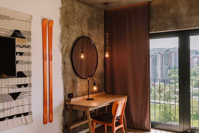 wnętrze pokoju ibis Styles Sarajevo od MIXD mały drewniany stolik i krzesło obok pomarańczowych nart na ścianie