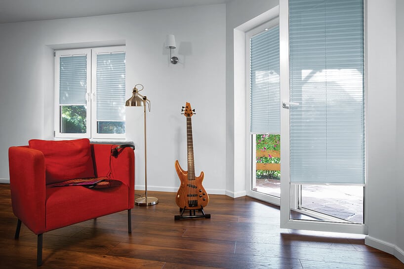 niebieskie plisowane żaluzje ANWIS w dwóch oknach w białym salonie z drewnianą podłogą i gitara na stojaku obok czerwonego fotela